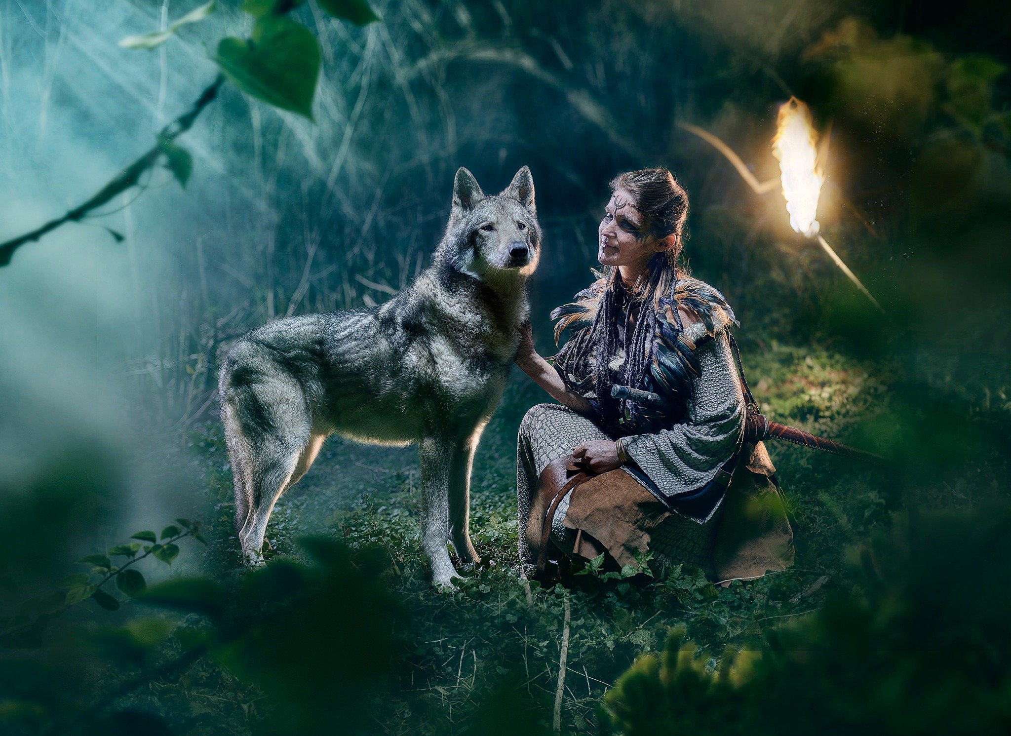 Une femme vêtue de vêtements médiévaux et tenant une épée, est agenouillée dans une forêt, caressant un loup gris.