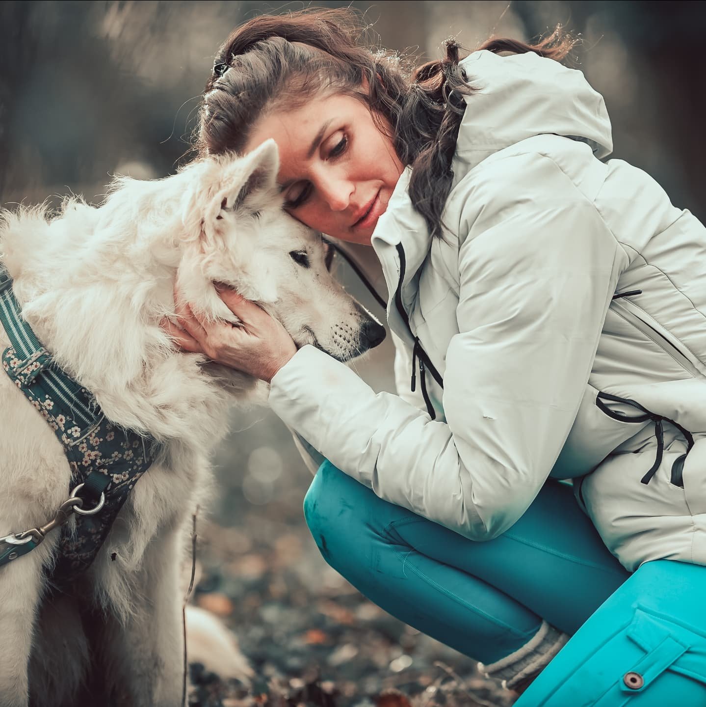 Une femme en extérieur, portant une veste blanche et un pantalon turquoise, caresse un chien blanc de grande taille avec un collier et un manteau, dans un environnement naturel, probablement en forêt.