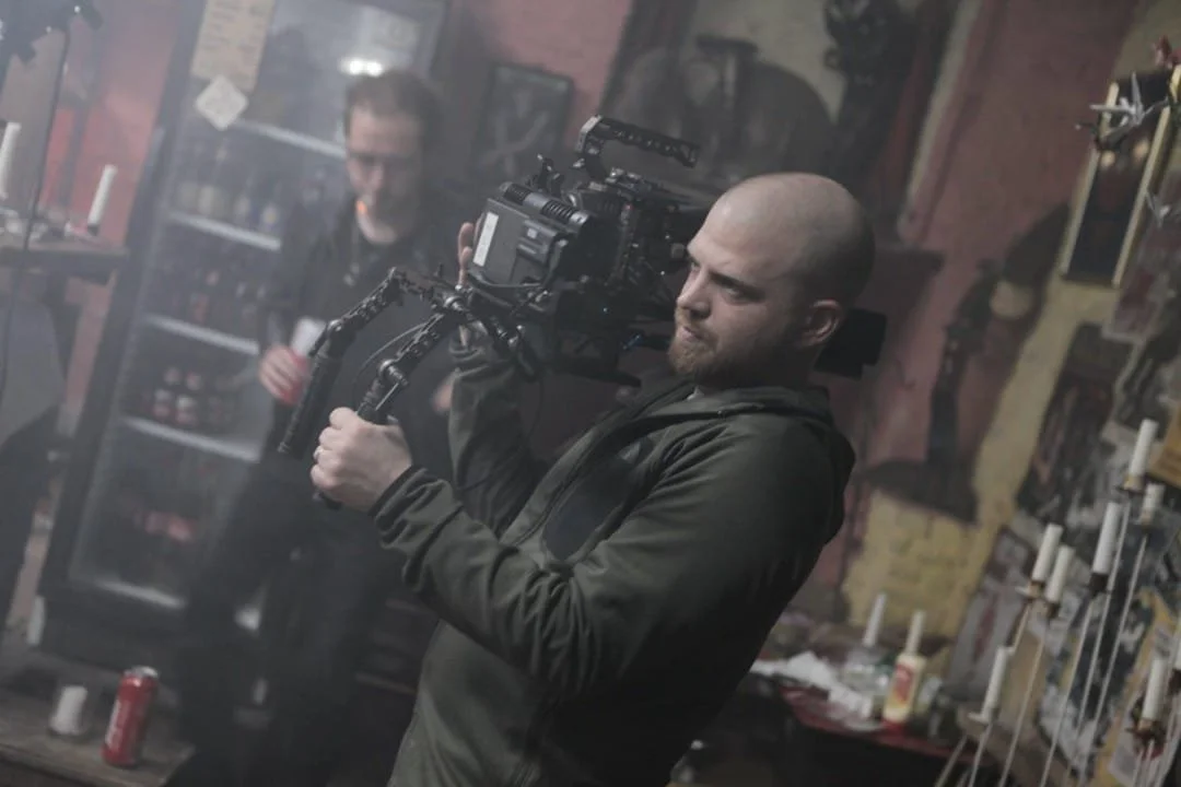 Un homme regarde à travers la poignée d'une caméra professionnelle dans un environnement intérieur, avec un autre homme flou en arrière-plan et diverses décorations sur les murs.