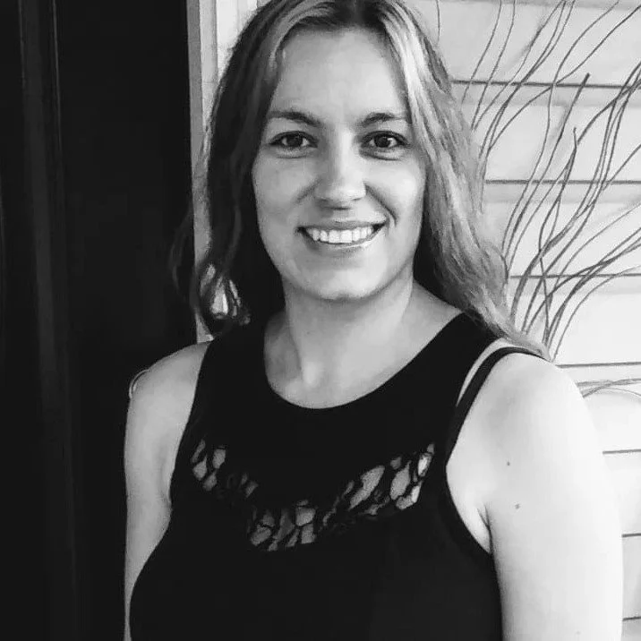 Black and white photo of a young woman smiling, wearing a sleeveless top with lace details, standing indoors with a patterned wall behind her.