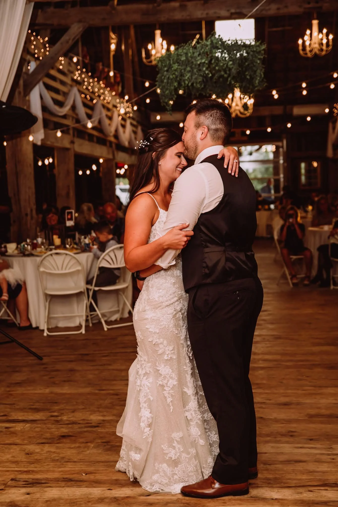 A bride and groom sharing a dance at their wedding reception in a rustic barn venue with warm lighting and hanging chandeliers.