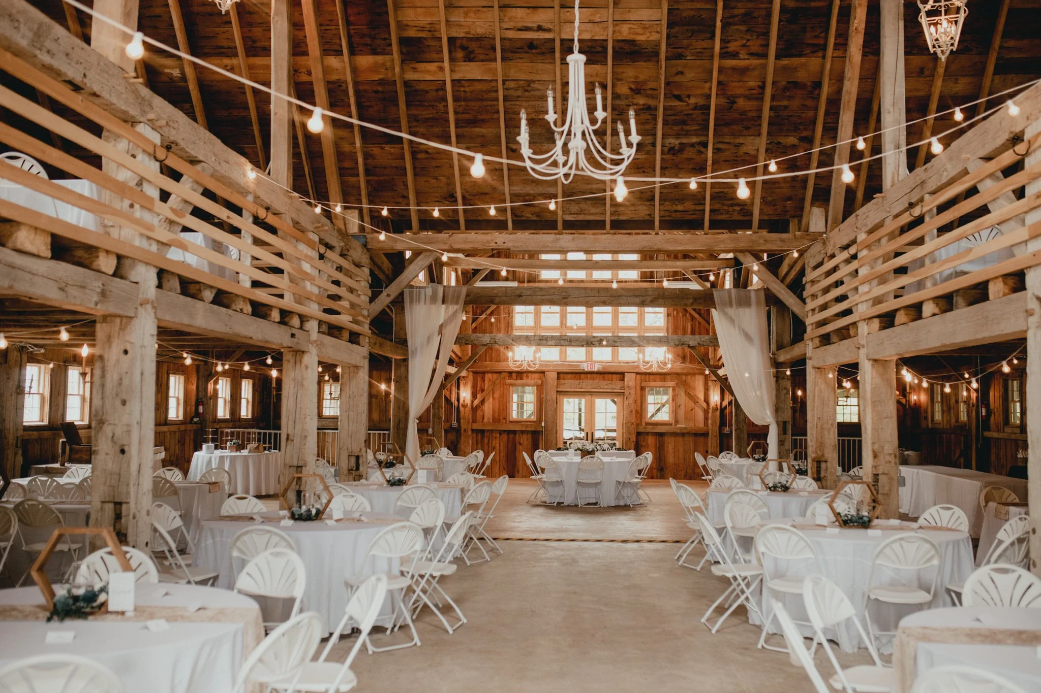 Interior of a rustic barn decorated for a wedding or event, with round tables covered in white tablecloths and white chairs. String lights hang from the wooden ceiling beams, and a large white chandelier is suspended in the center. Natural light filters through multiple windows.