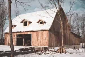Snow-covered rustic barn with a pitched roof in a winter landscape, surrounded by leafless trees.