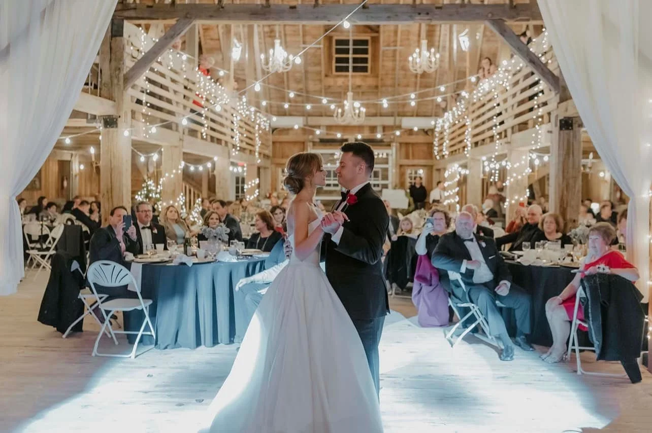Bride and groom sharing first dance at wedding reception in rustic barn decorated with string lights and chandeliers, guests seated at round tables watching.