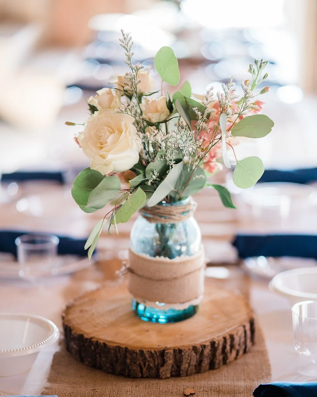 A floral centerpiece with white roses, pink flowers, and greenery in a blue glass jar decorated with burlap, set on a wooden slab at a table.