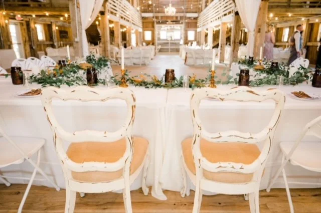 Wedding reception decorated with white tablecloths, floral centerpieces, and vintage chairs, set in a rustic barn with warm lighting and white drapes.