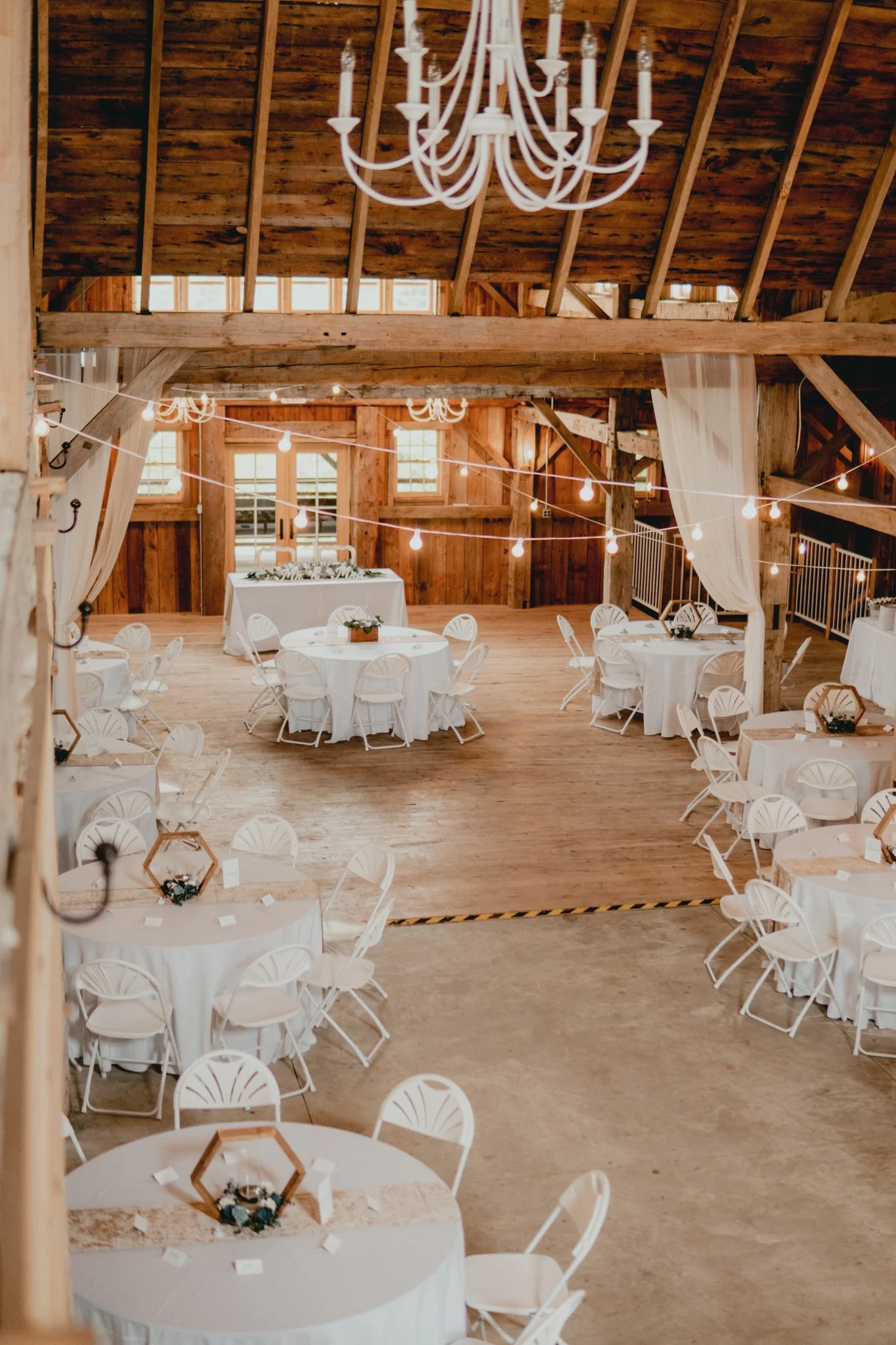 Interior of a rustic wooden barn decorated for an event with round tables covered in white tablecloths and white chairs, string lights hanging from the ceiling, and a large white chandelier.