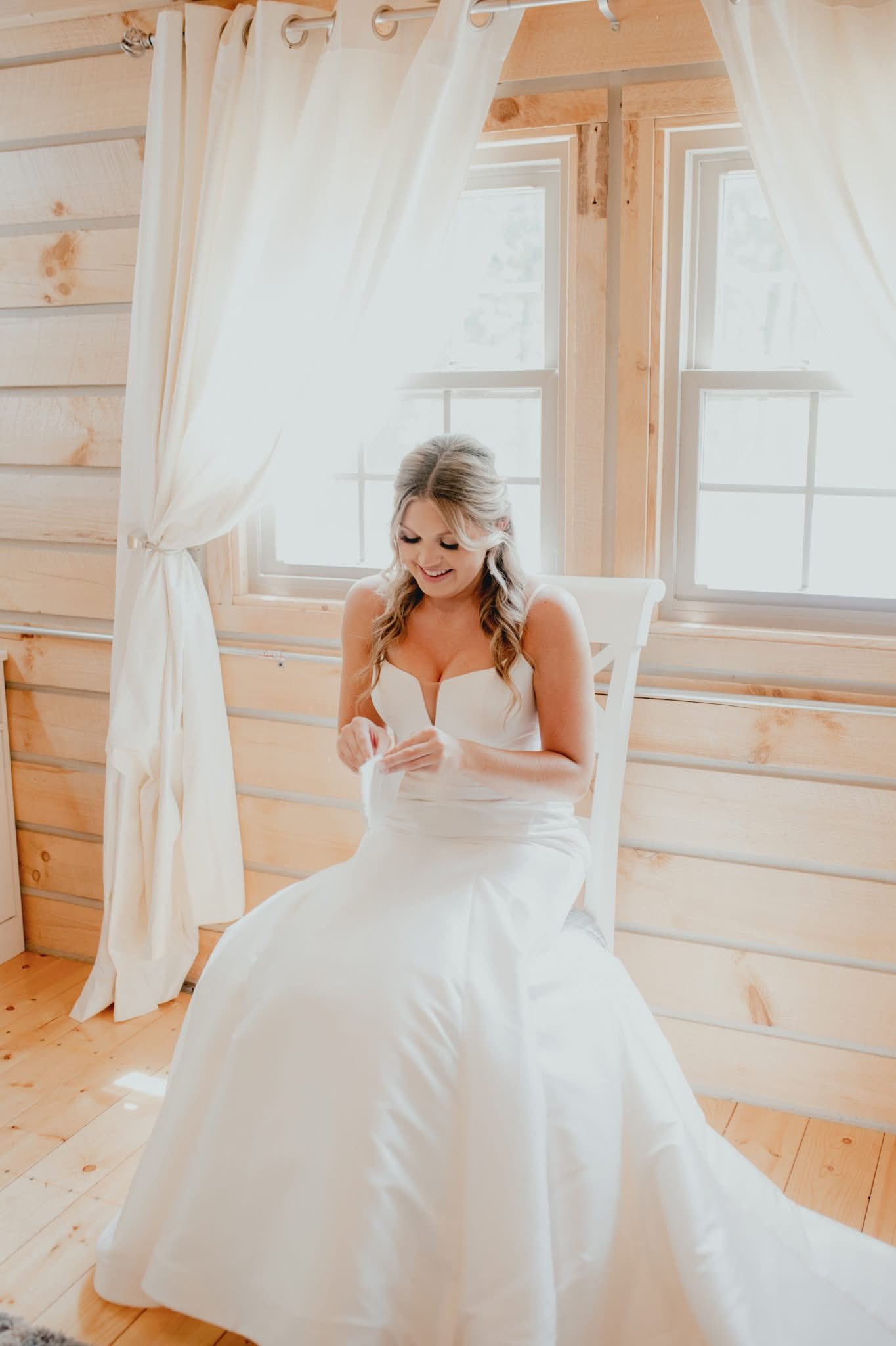 A woman wearing a white wedding dress sitting on a white wooden chair inside a sunlit room with wooden walls and large windows, smiling and looking down.