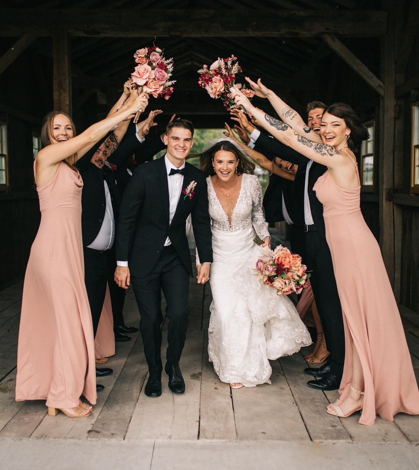 A bride and groom walking hand-in-hand under a wedding arch with friends celebrating around them, holding flower bouquets in a rustic wooden venue.