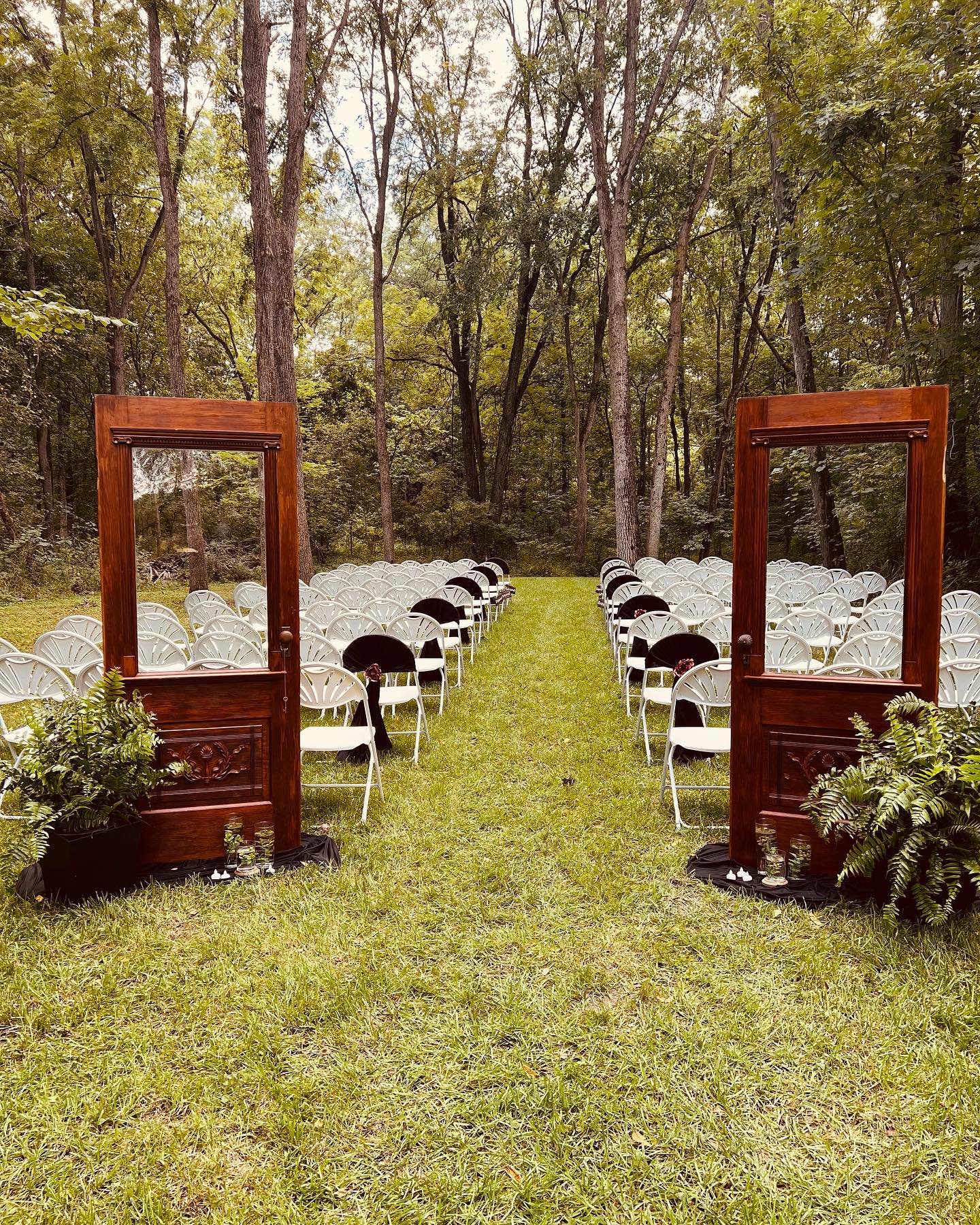 Outdoor wedding ceremony setup with rows of white chairs, black chairs, and wooden frame doors on a grassy area surrounded by trees.