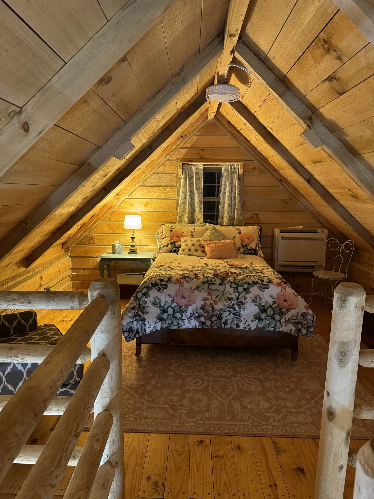 A cozy bedroom with wood-paneled sloped ceiling, a window with floral curtains, a floral bedspread, a nightstand with a lamp, a small air conditioner, and a chair.