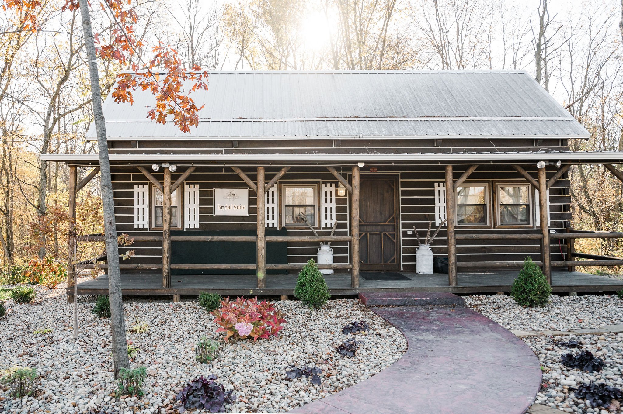 A rustic wooden building with a sign that reads 'Bridal Suite', set in a wooded area with trees, a small garden with flowers and bushes, and a gravel pathway leading to the entrance.