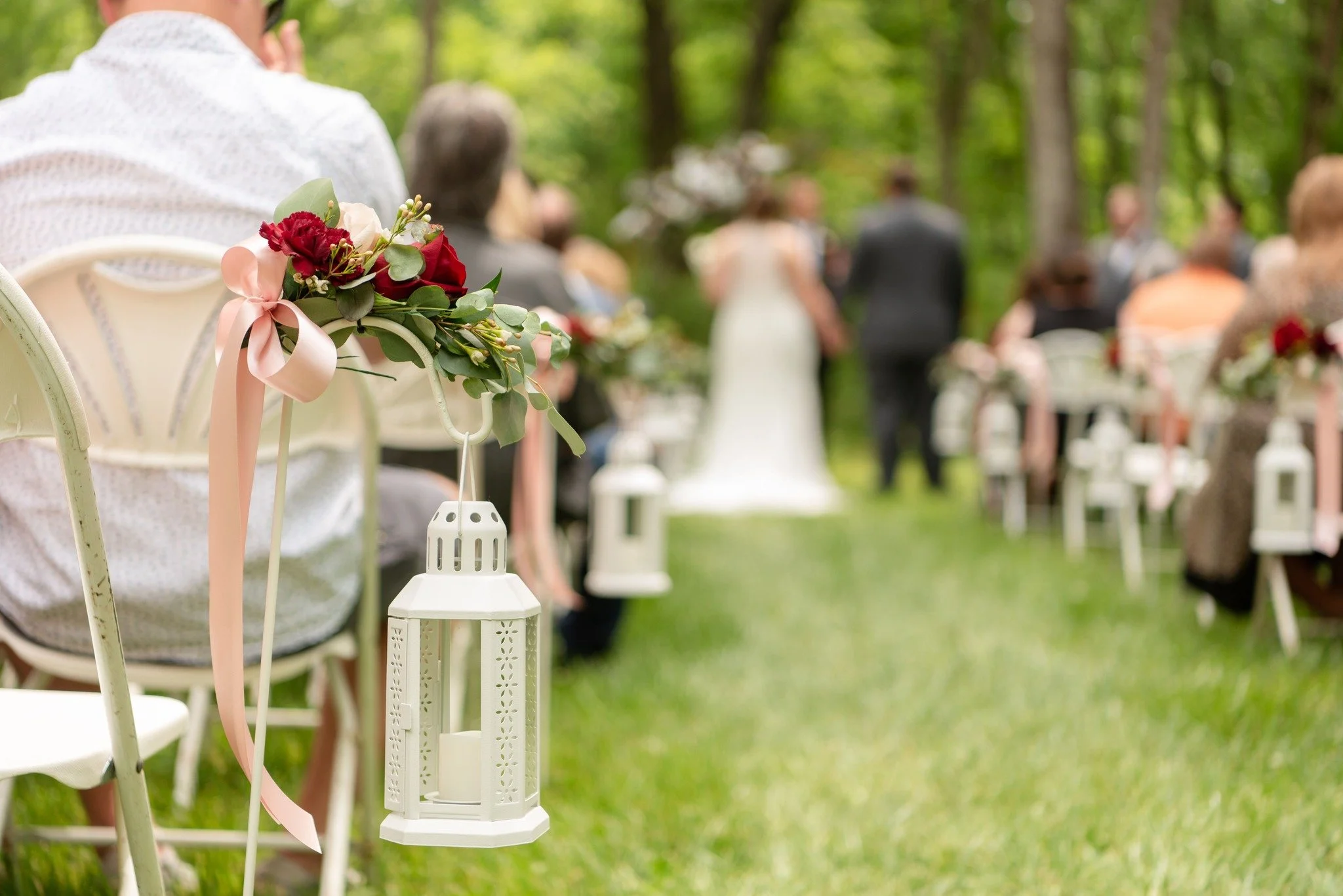 Decorative chairs with floral arrangements and hanging lanterns set up outdoors in a wooded area for a wedding or outdoor event.