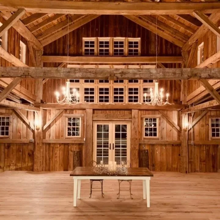 Interior of a rustic wooden barn with high ceiling, chandeliers, and a farmhouse table with chairs.