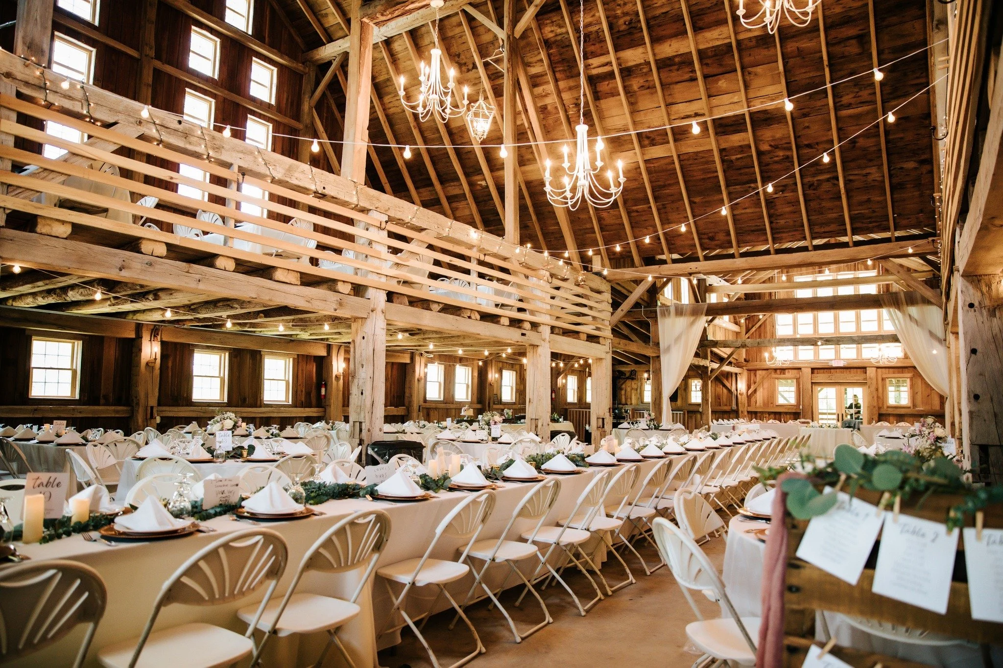 Rustic banquet hall decorated with string lights and chandeliers, set up for a wedding reception with rows of white chairs and tables adorned with floral centerpieces and place settings.