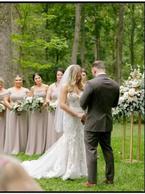 Bride and groom holding hands during a wedding ceremony in a forest, with bridesmaids standing behind them holding bouquets.