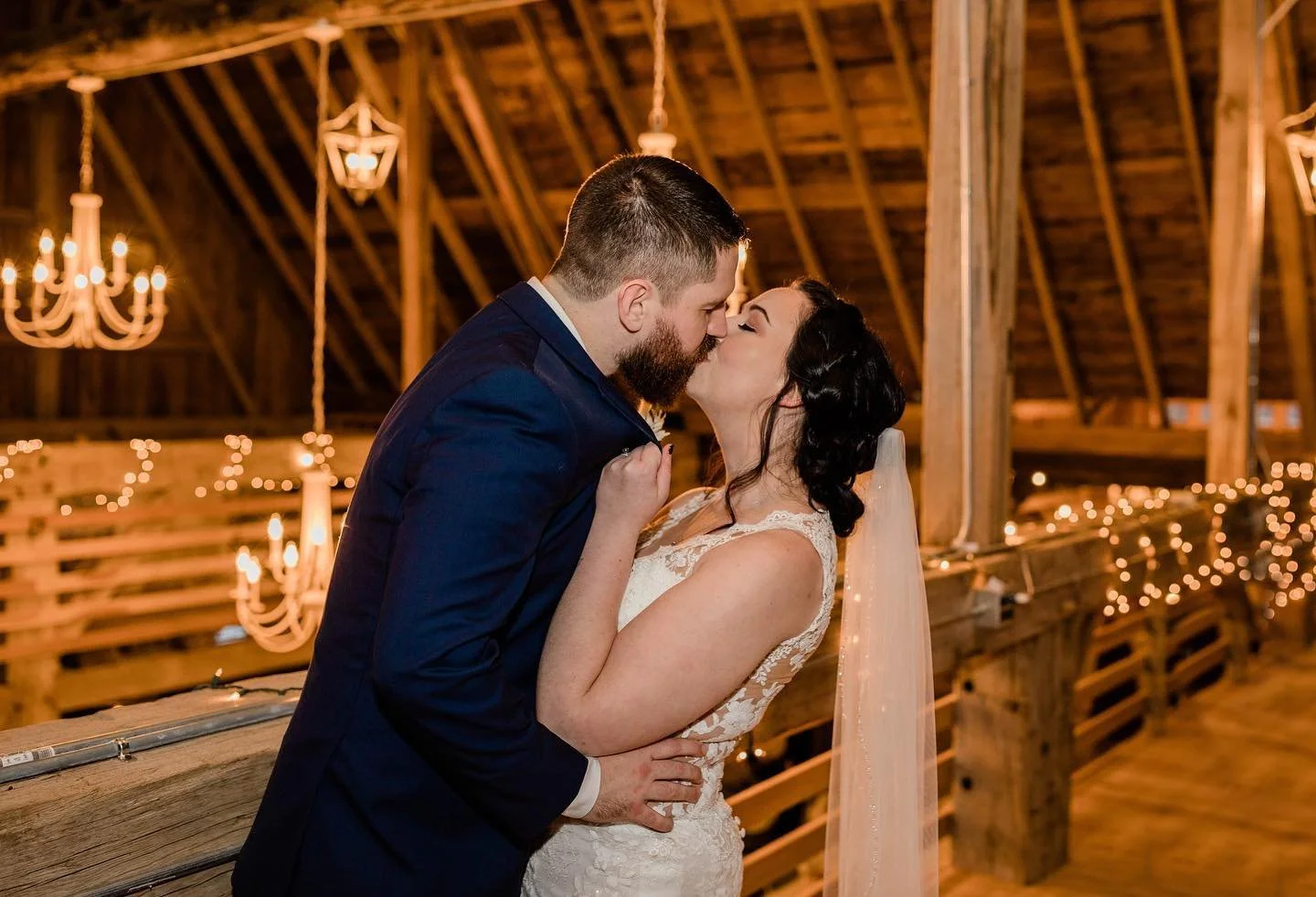 A bride and groom kiss in a rustic barn decorated with string lights and chandeliers during their wedding reception.