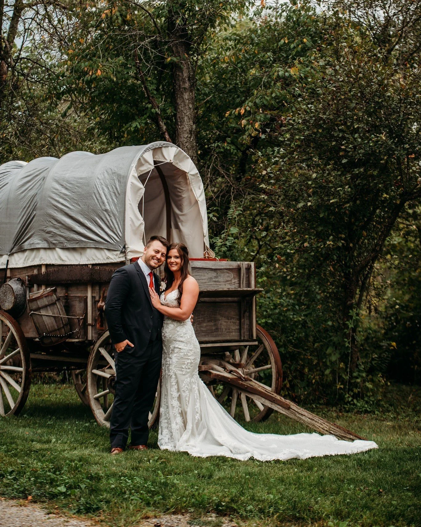 A bride and groom stand together in front of a wooden covered wagon with a fabric canopy, surrounded by trees with green leaves. The bride is in a white lace wedding dress, and the groom is in a dark suit with a red tie. They are smiling and holding hands, celebrating their wedding day outdoors.