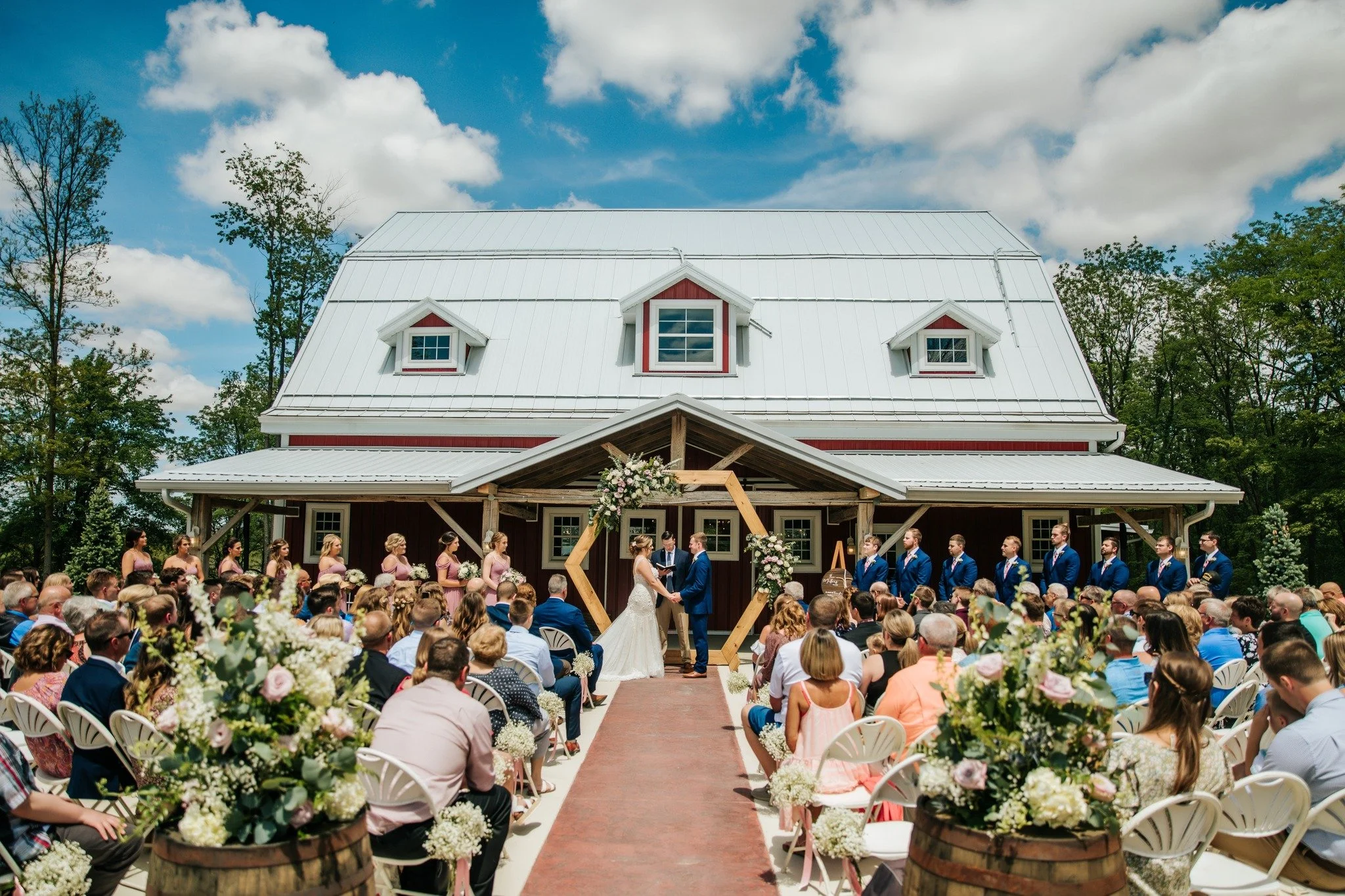 A wedding ceremony taking place outdoors in front of a rustic barn with a crowd of guests. The bride and groom are exchanging vows under a wooden arch decorated with flowers.