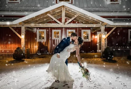 A newlywed couple dancing outside on snow in front of a barn with string lights.