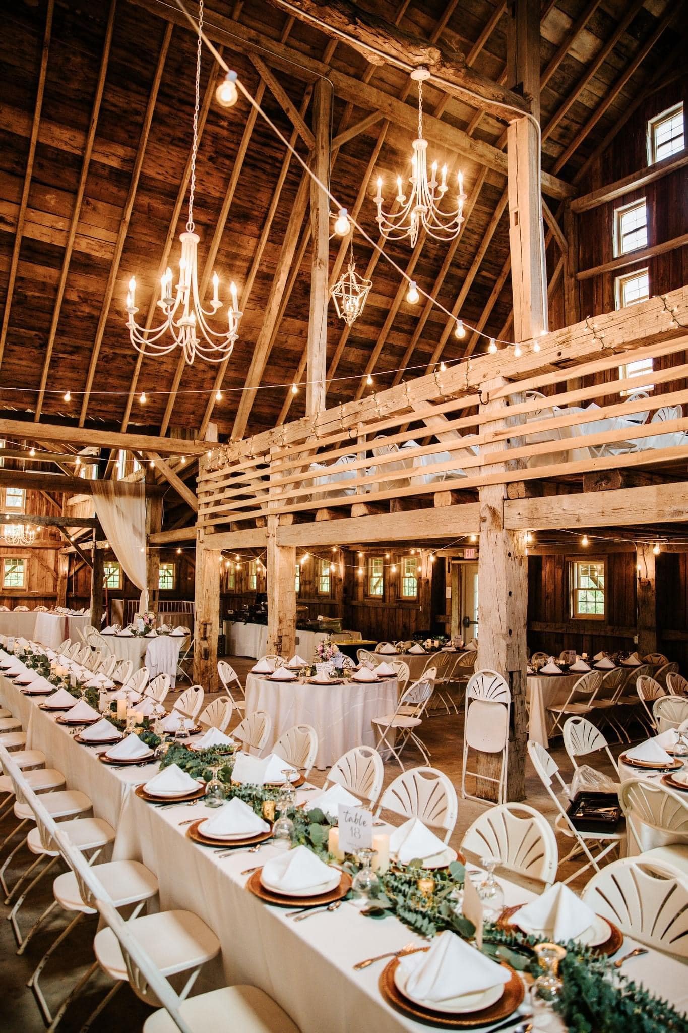 Long banquet tables, white chairs, greenery, candles, and elegant chandeliers hanging from the wooden ceiling adorn the rustic barn reception area.