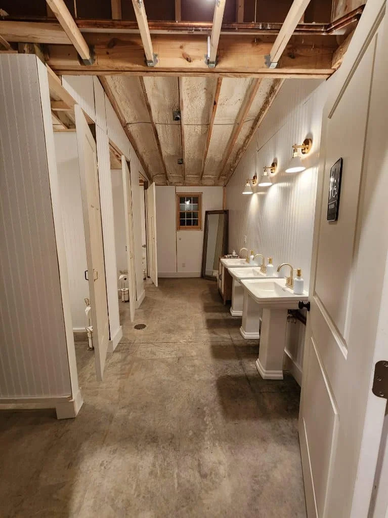 Interior view of a rustic bathroom under renovation with three sinks, gold fixtures, mirror, and unfinished ceiling showing wooden beams.