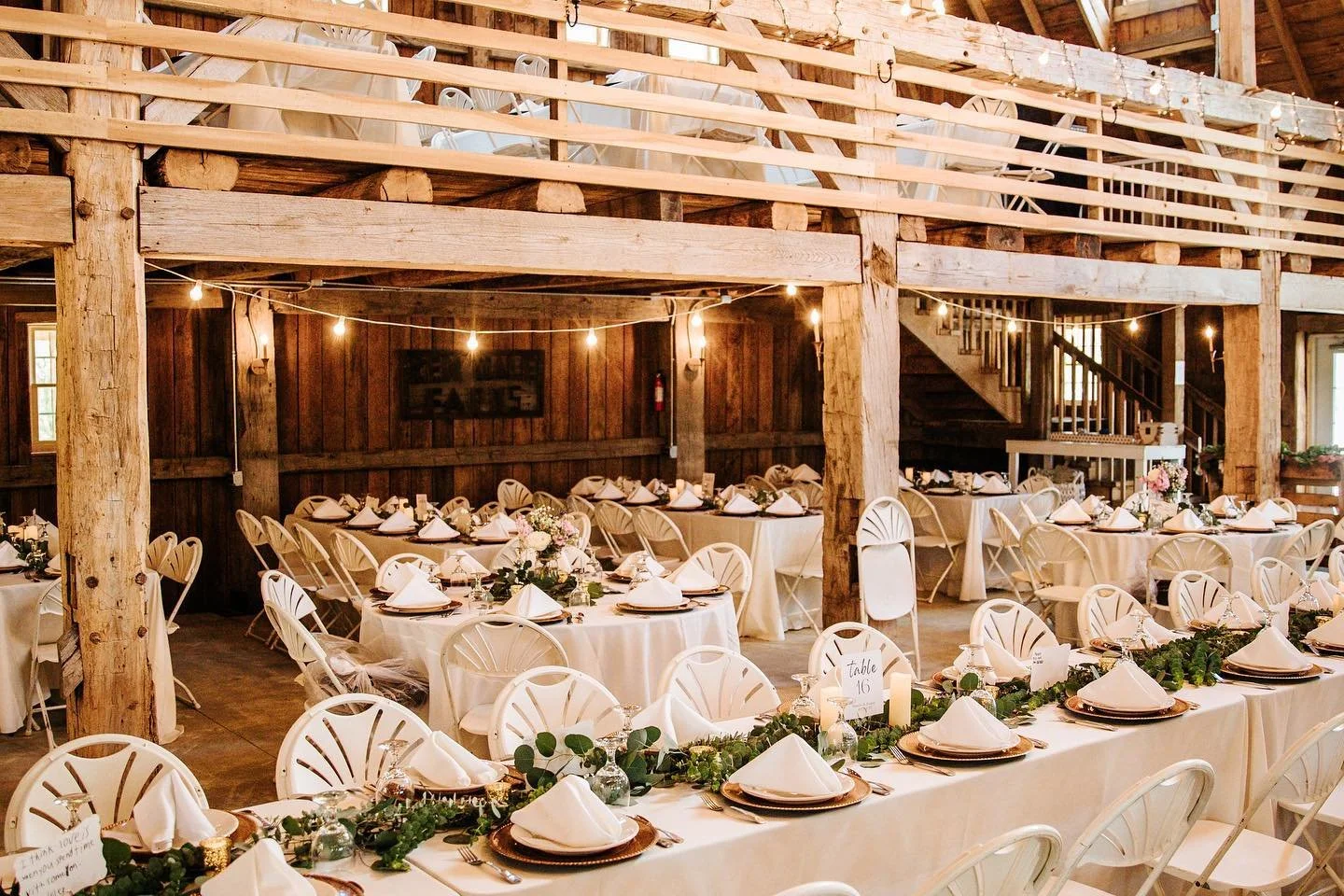 Rustic wedding reception with round and rectangular tables decorated with white tablecloths, white napkins, greenery, and pink flowers. The space features exposed wooden beams, string lights, and wooden railing on second level.