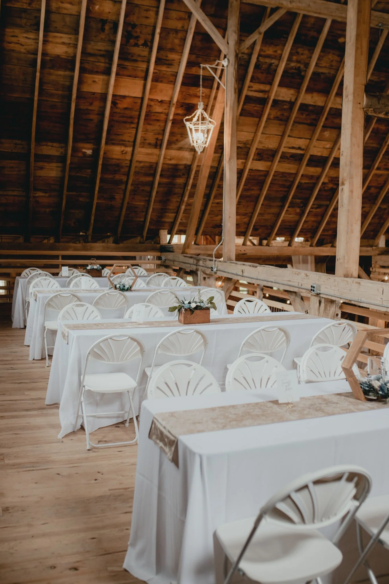 Wedding or event reception setup inside a rustic barn with wooden beams and a sloped ceiling. Long tables covered with white tablecloths and decorated with floral centerpieces are arranged. White chairs are placed around the tables. A chandelier hangs from the ceiling.