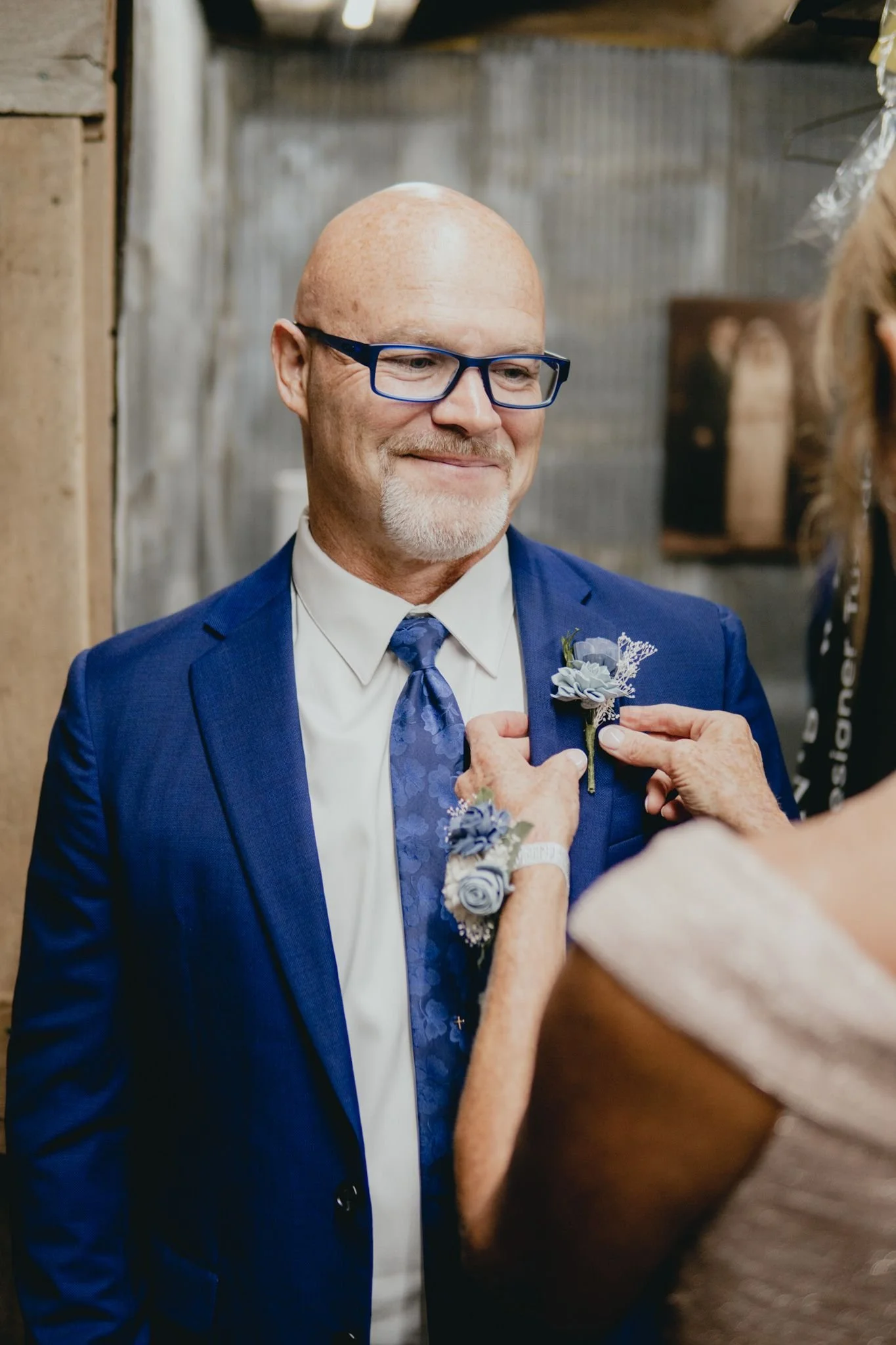 A man in a blue suit with glasses and a boutonnière on his lapel smiling as a woman pins a boutonnière on his suit at a wedding celebration.