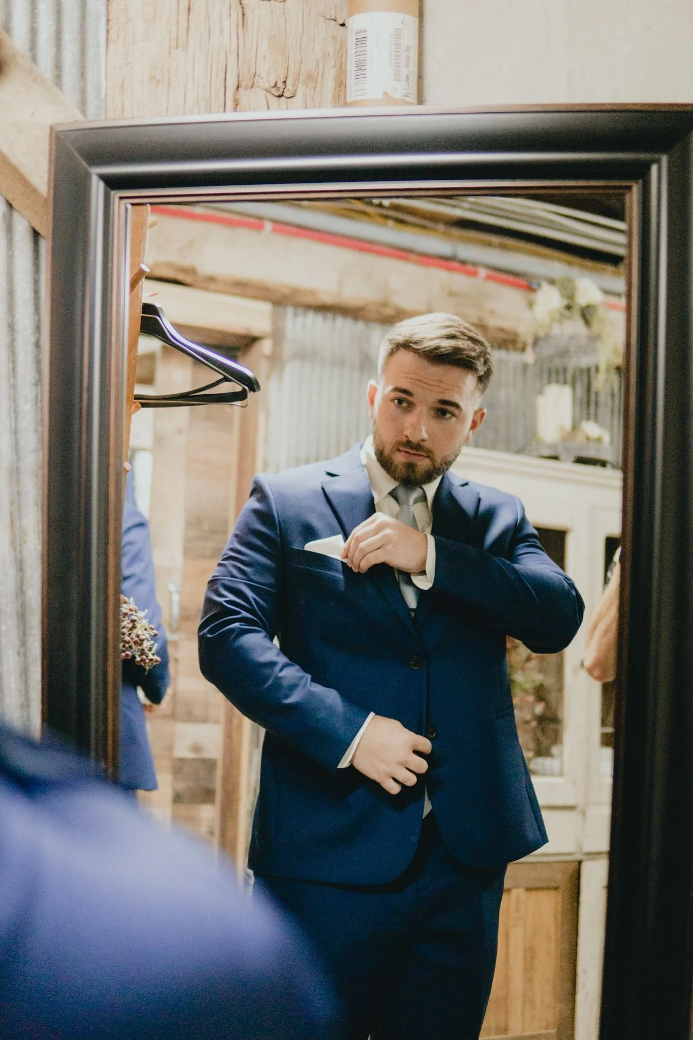 A man in a blue suit and tie is adjusting his cuff in front of a mirror, with wooden walls and hanging hangers in the background.