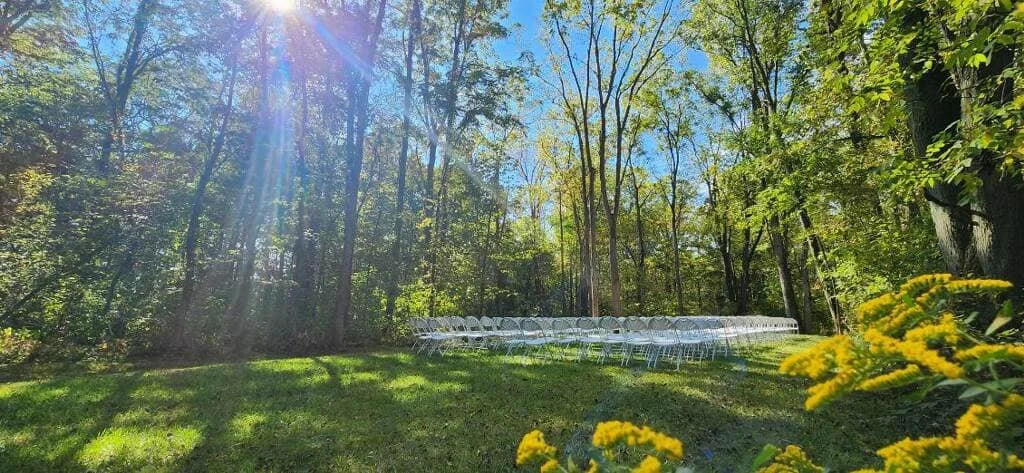 Outdoor gathering area with a circle of white chairs on a grassy lawn, surrounded by trees, under a bright blue sky with sunlight streaming through the trees.