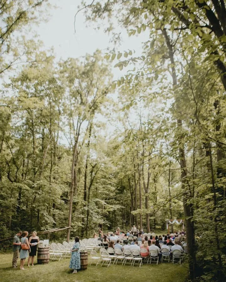 Outdoor wedding ceremony seating in a forest clearing with chairs arranged in rows, some guests seated, standing guests, and a small arch decorated with flowers.