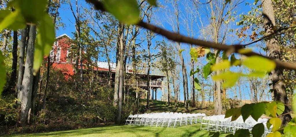 An outdoor wedding setup with white chairs arranged on green grass, set against a backdrop of trees and a house elevated on stilts under a clear blue sky.