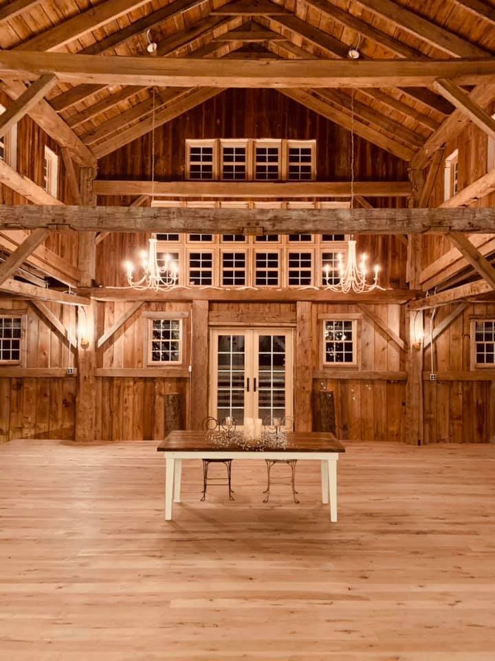 Spacious wooden barn interior with high vaulted ceiling, multiple windows, and a central dining table. Chandeliers and wall sconces provide lighting.