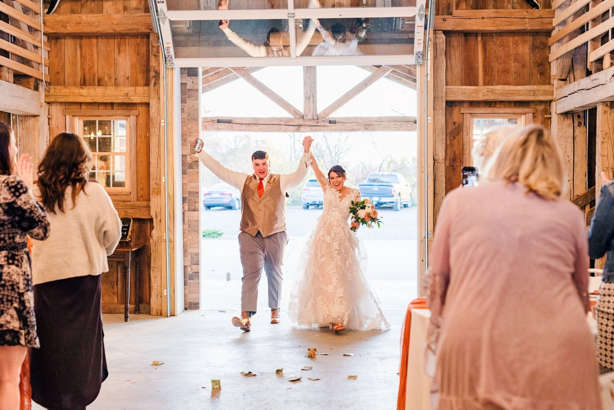 A newlywed couple celebrating as they walk through a rustic barn door into a wedding reception, with guests standing and clapping on either side.