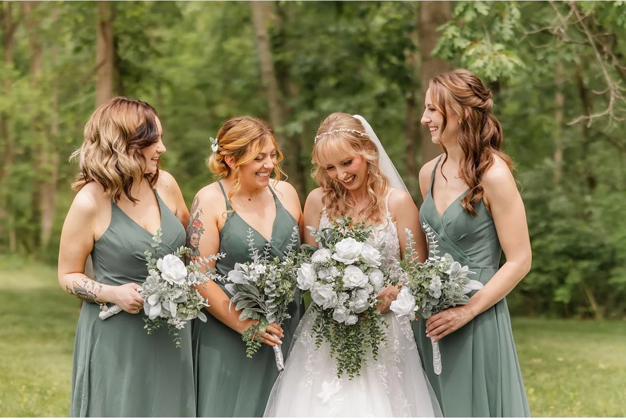 Bride and four bridesmaids standing outdoors in a wooded area, all holding bouquets of white flowers and greenery, smiling and looking at each other.