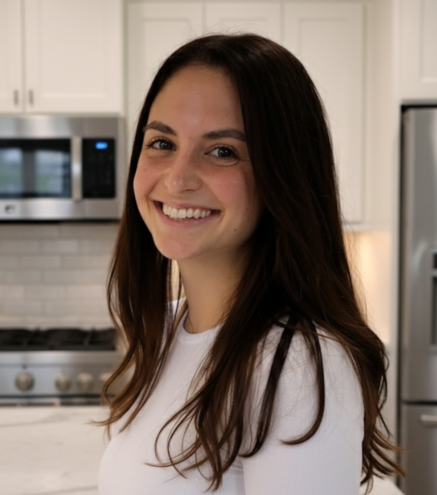 A woman with long brown hair smiling in a kitchen with white cabinets and stainless steel appliances.