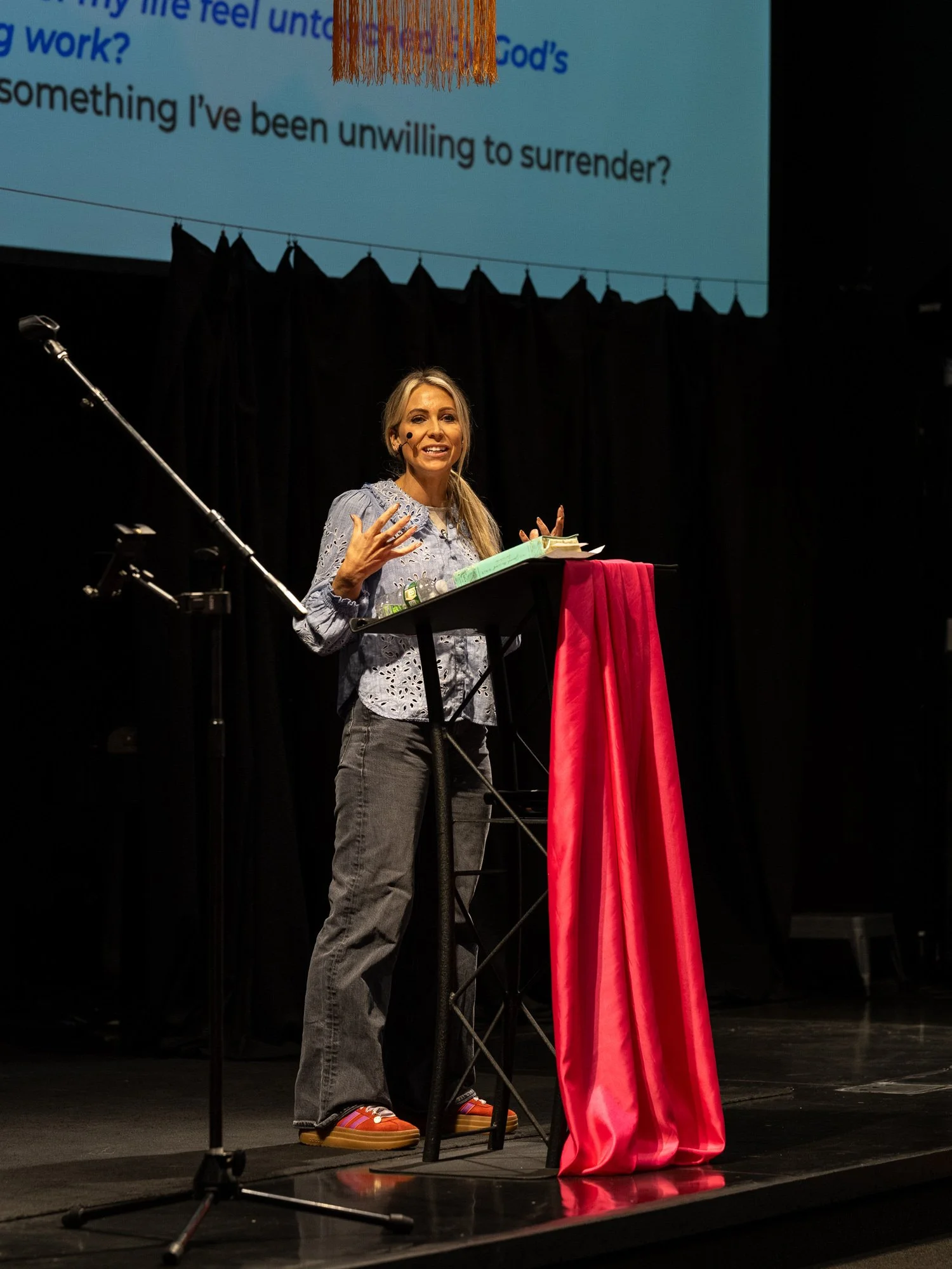 A woman stands behind a lectern on a stage, speaking into a microphone. The stage has a black curtain backdrop, and a large screen above displays text. The lectern is covered with a pink cloth. The woman has blonde hair, is wearing a gray patterned blouse, gray jeans, and colorful shoes, and appears to be engaged in speaking or teaching.