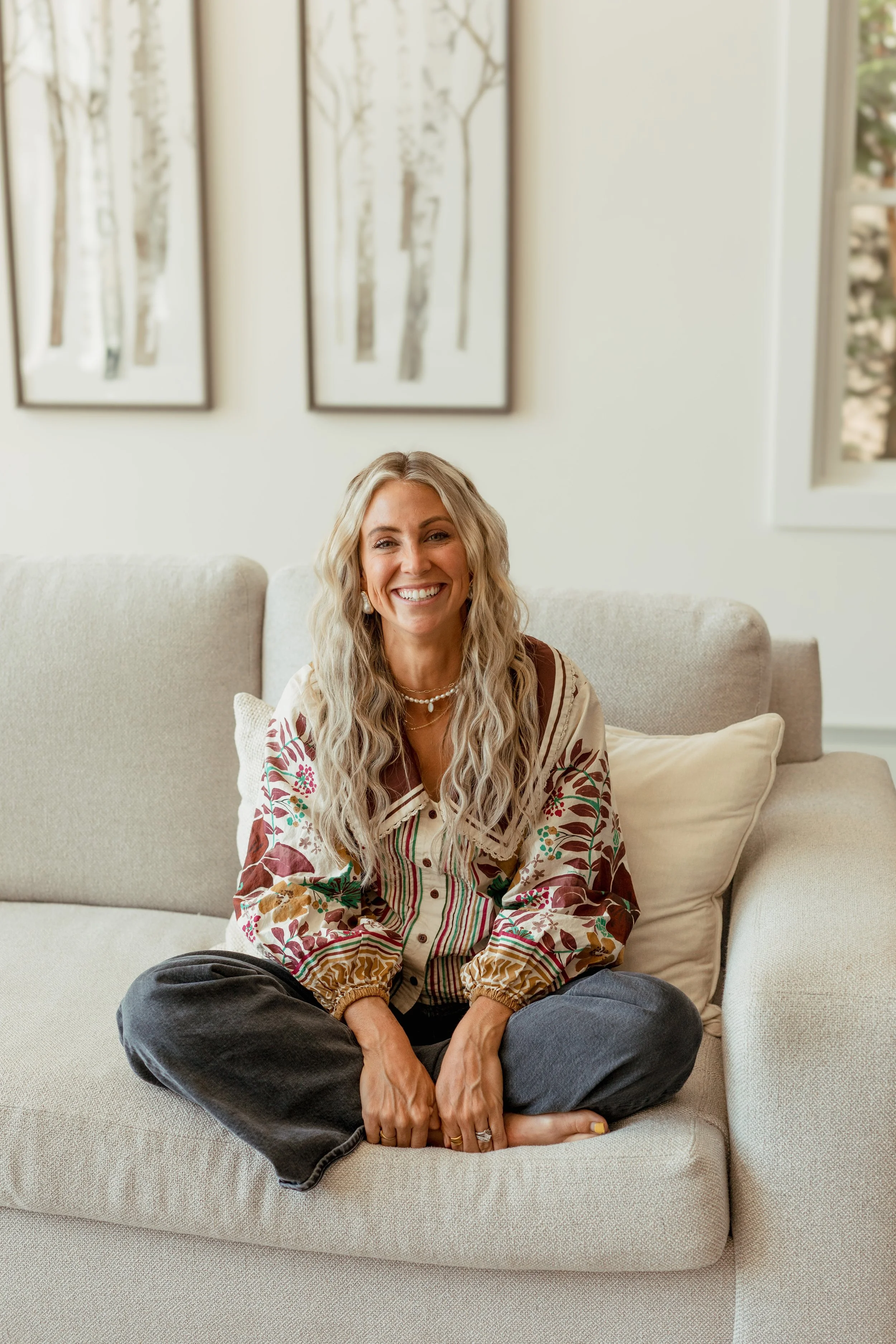 A woman with long, wavy blonde hair sitting cross-legged on a light-colored sofa, smiling at the camera. She is wearing a floral-patterned shirt, dark jeans, and layered necklaces. Behind her are two framed pictures of trees and a window with greenery outside.