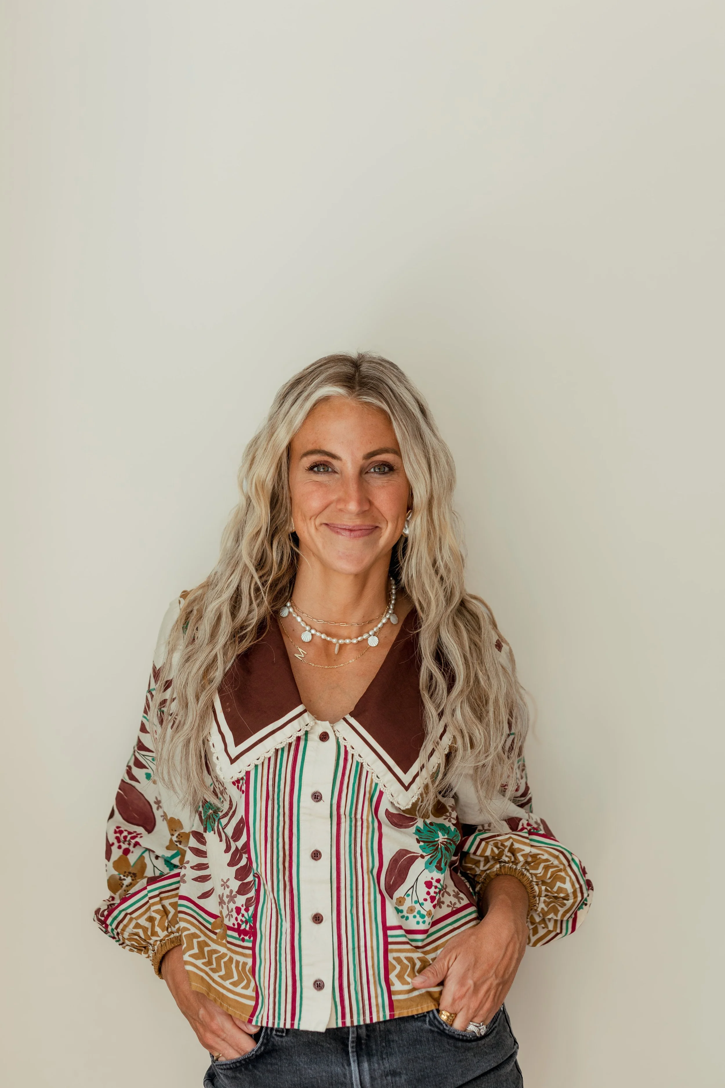 A woman with long, curly blonde hair wearing a colorful patterned shirt and layered jewelry, smiling against a plain white wall.