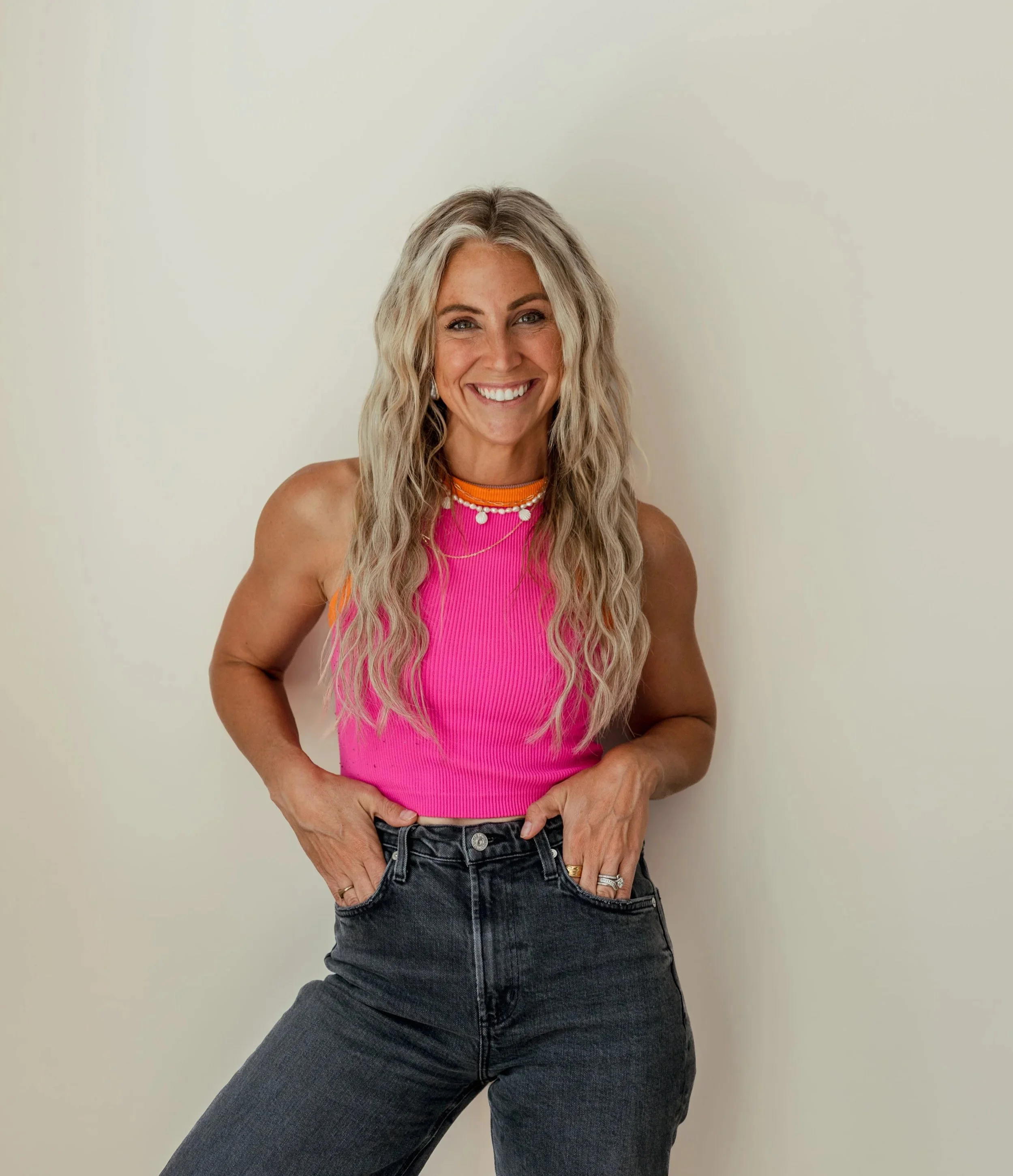 A woman with long blonde wavy hair smiling, wearing a bright pink sleeveless top and dark jeans, standing against a plain light background.