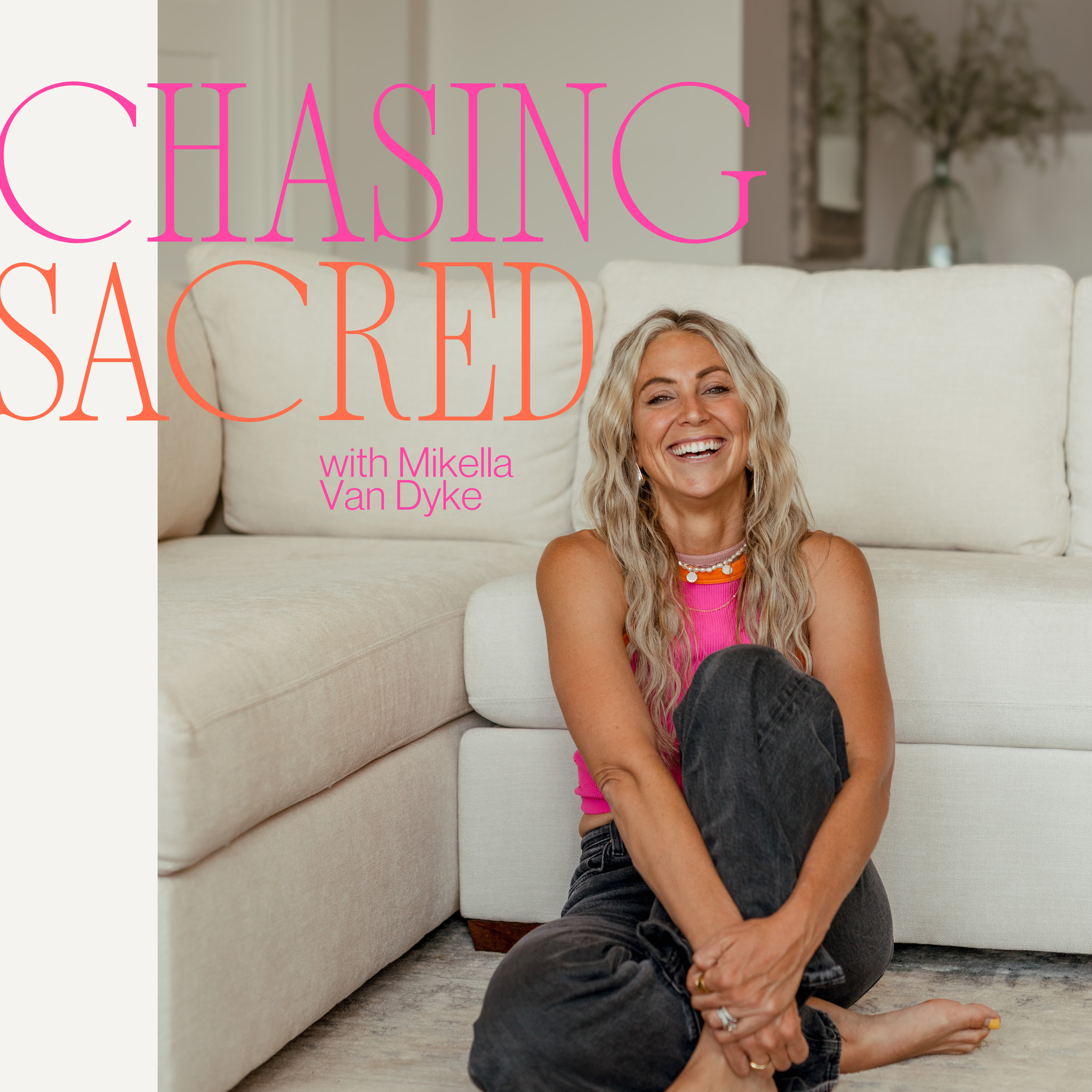 A woman with long wavy blonde hair sitting on the floor in front of a cream-colored sofa, smiling with her legs crossed and wearing a pink sleeveless top and dark jeans, with decorative text overlay reading 'Chasing Sacred with Mikella Van Dyke.'