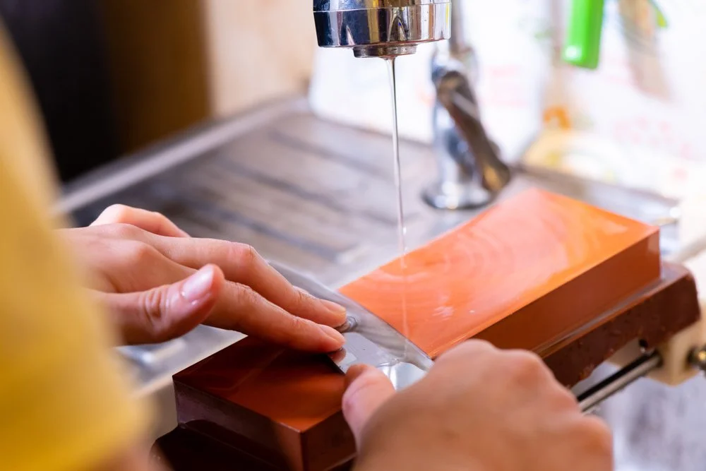 Someone sharpening a knife on a wet stone with water flowing on it in a kitchen.