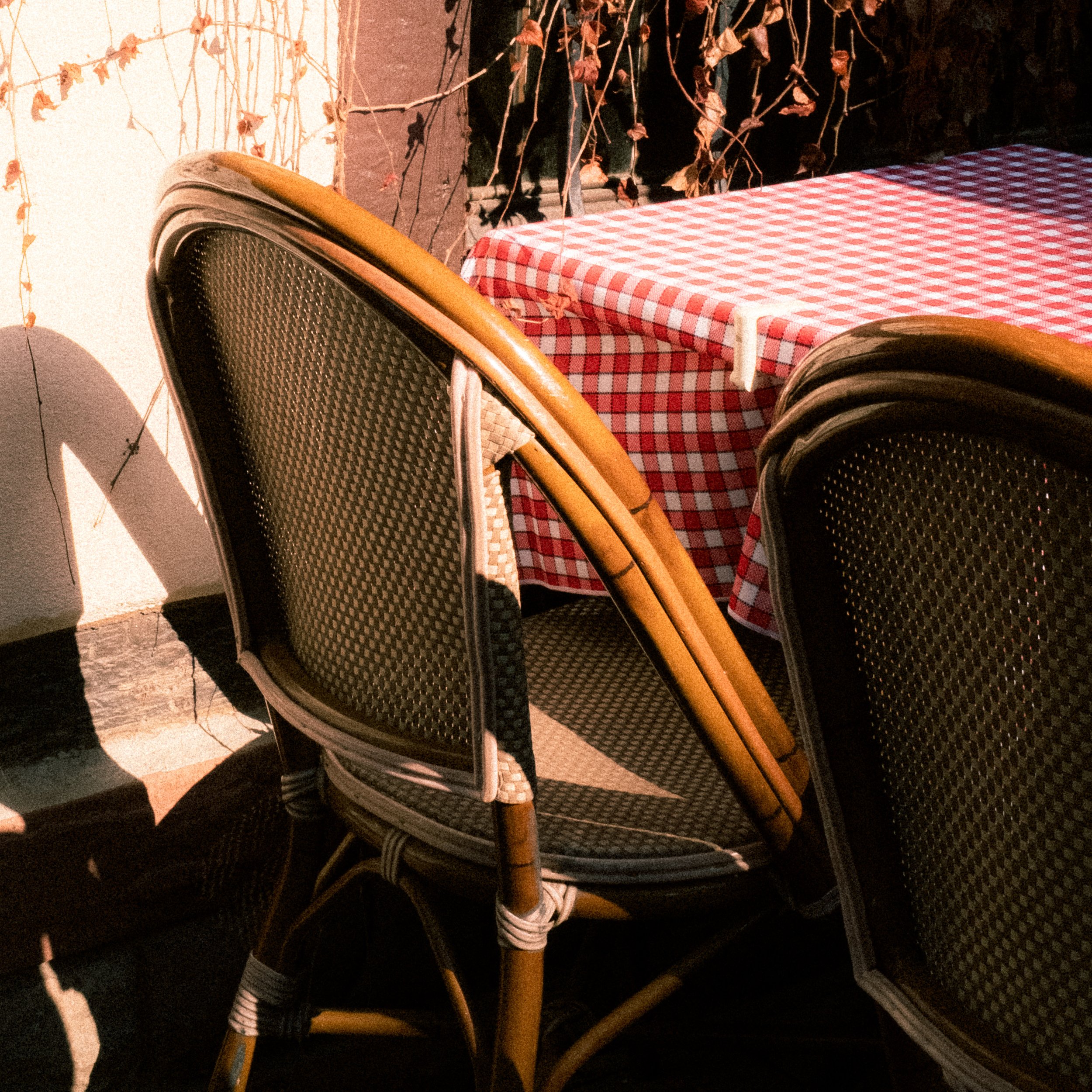 Chaises en rotin empilées près d'une table à couverture à carreaux rouges et blancs, en plein soleil, avec un mur blanc et des plantes sèches en arrière-plan.