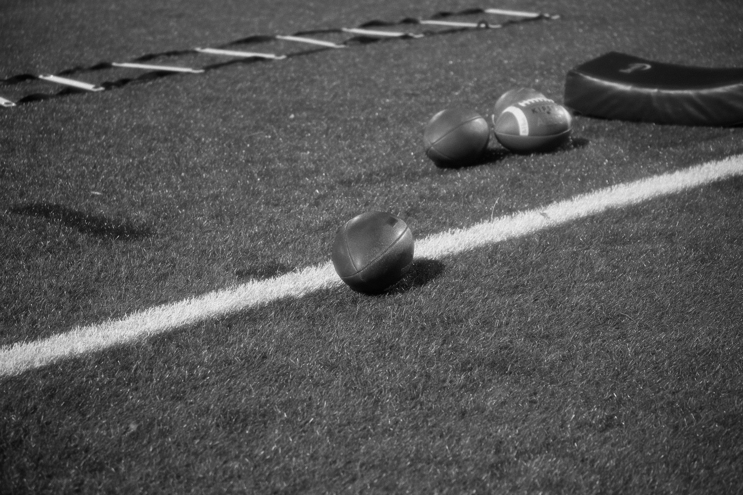 Une ligne de différents types de balles de football américain et de matériel de musculation placés sur un terrain, avec des outils d'entraînement visibles au fond.