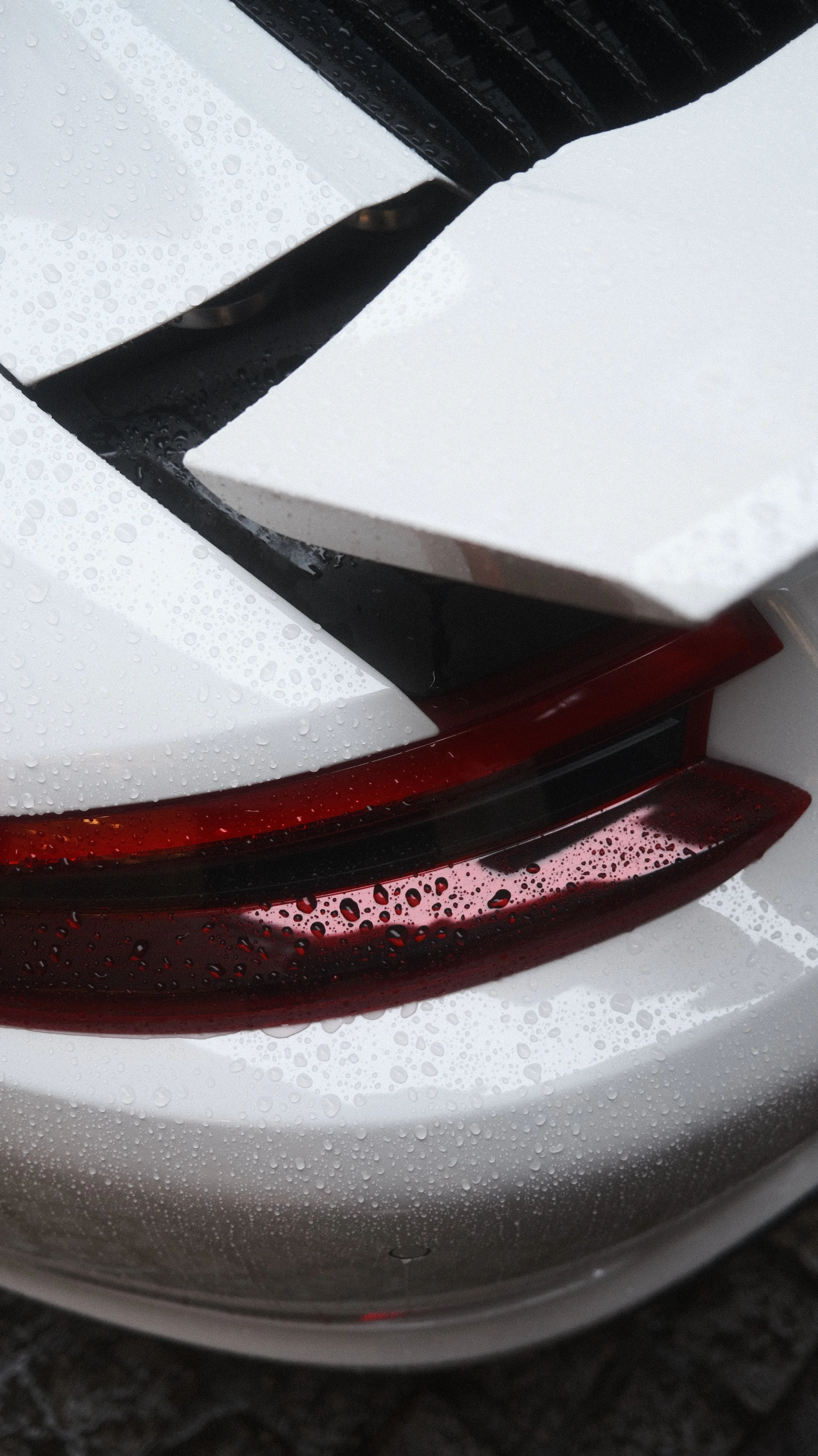 Close-up de la partie arrière d'une voiture blanche, montrant la lumière arrière rouge, des gouttelettes d'eau sur la surface, avec un aileron noir partiellement visible en haut.