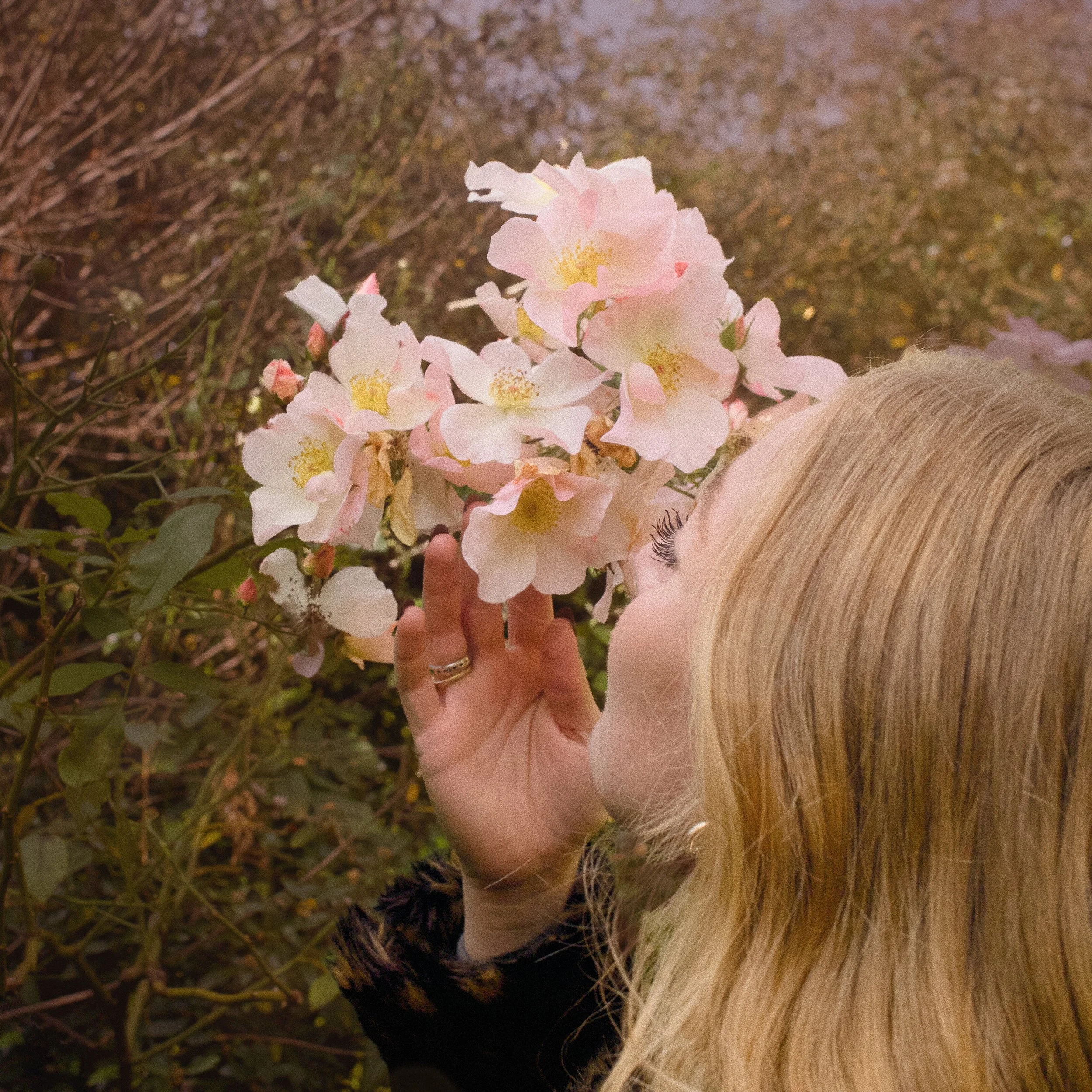 Femme avec cheveux blonds tenant un bouquet de fleurs roses et blanches dans un environnement naturel.