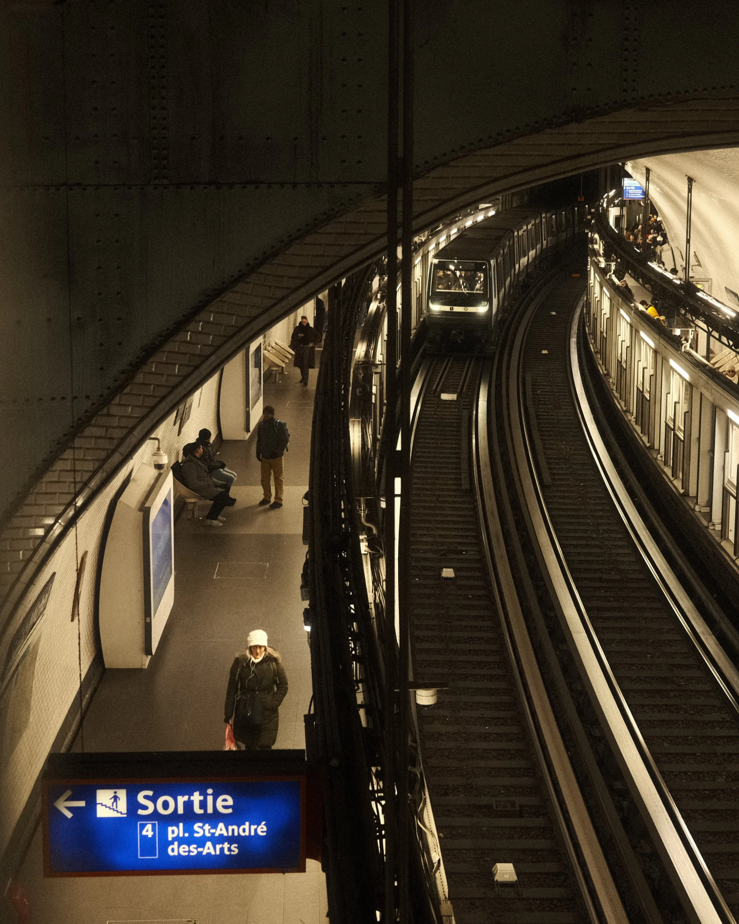 Une station de métro avec des passagers, un train arrivant sur la voie, et un panneau indiquant la sortie vers la place Saint-André des Arts.