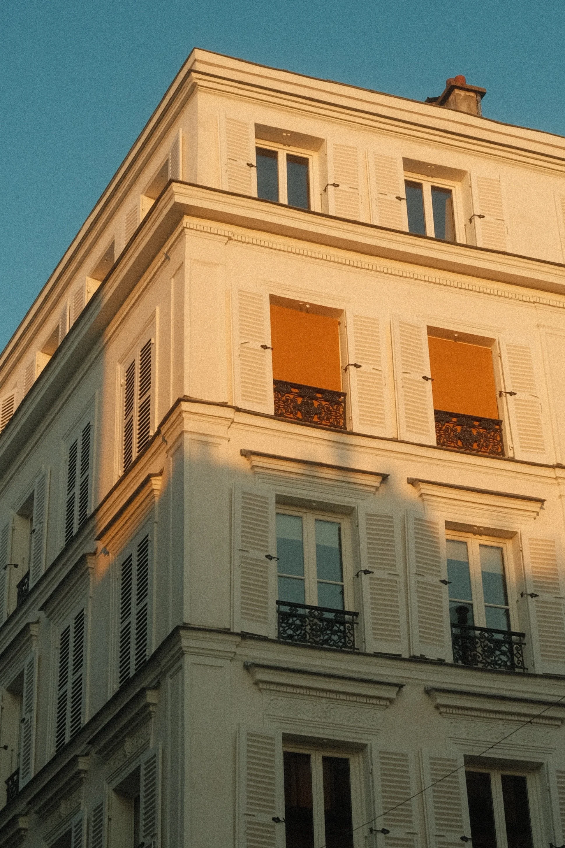 Un bâtiment résidentiel à plusieurs étages avec des fenêtres dotées de volets blancs et de balcons en fer forgé, éclairé par la lumière du coucher du soleil.