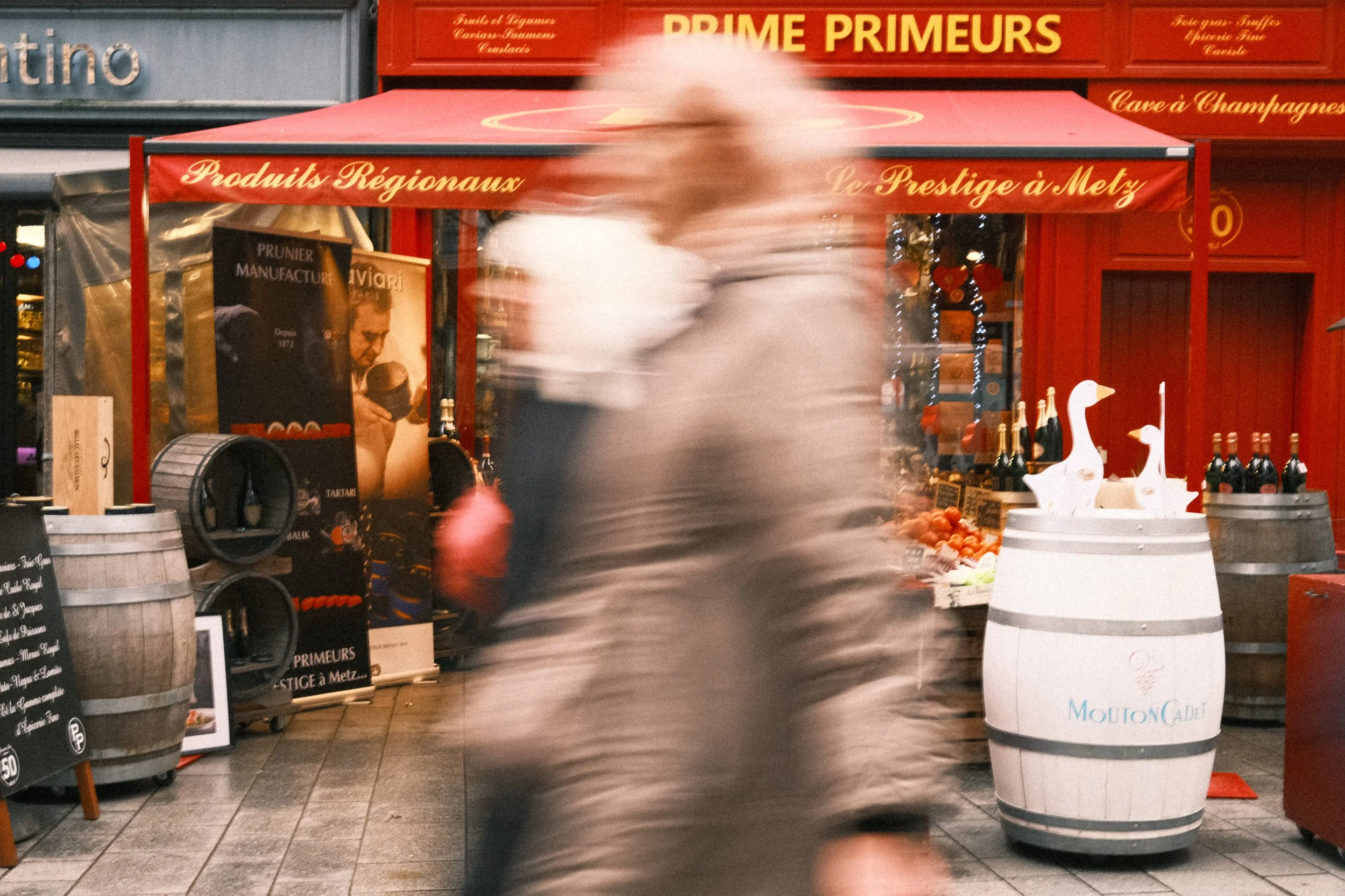 Une scène urbaine avec une personne floue marchant devant un stand de marché ou de vente de produits régionaux, avec des bouteilles de champagne et des décorations en forme de canards en blanc, marqué 'Mouton Cadet'.